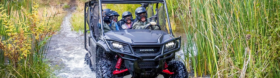 Four people riding in a 2018 Honda® Pioneer™ 1000-5 Deluxe side by side through a river canal near tall grass reeds.